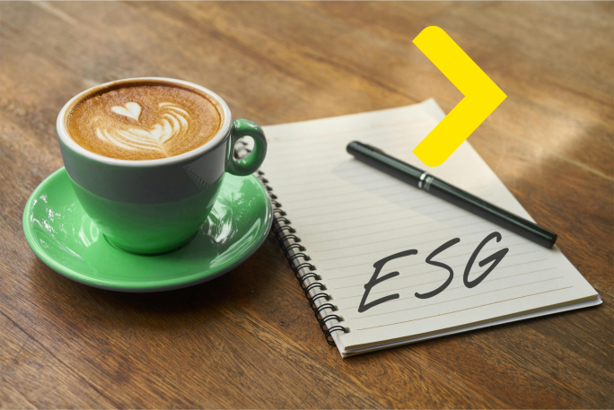 Зелена ащка з кавою-лате з латте-артом у формі серця на деревяному столі поруч із блокнотому із написаом ESG